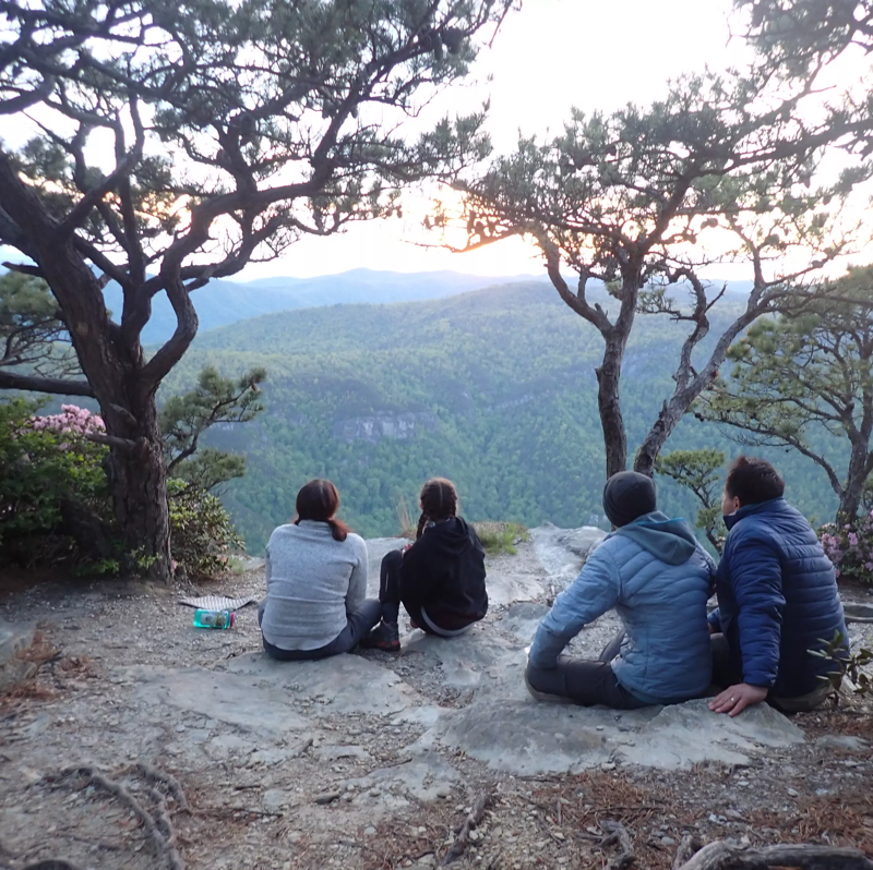 The image shows four people sitting on the edge of a cliff, looking out at a scenic view of mountains and trees. The sun is setting in the distance, casting a warm glow over the landscape. The people are dressed in casual clothing, and they appear to be enjoying the view. There are trees framing the image on both sides.